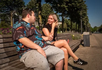 Een jongeman en een jonge vrouw zitten buiten pratend op een lange houten bank.